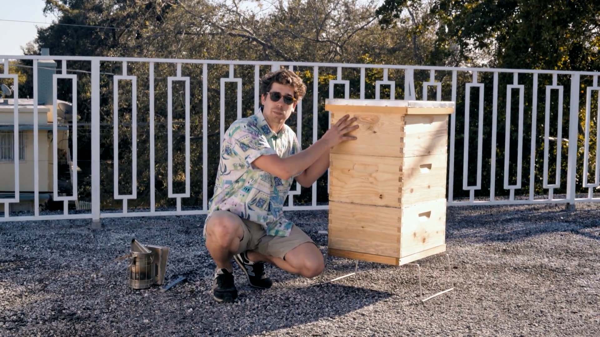 A beekeeper crouches beside a beehive, with a smoker at his side