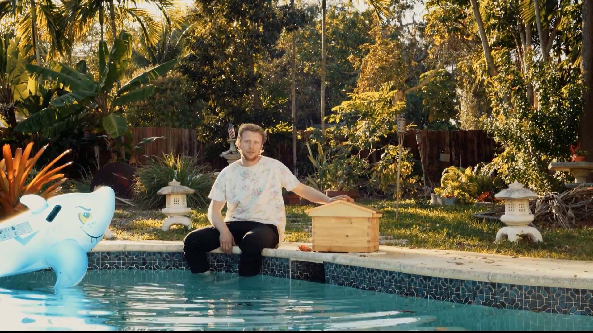 A beekeeper sits at the side of a pool with his feet in the water and one hand resting on a beehive. An inflatable shark floats beside him.