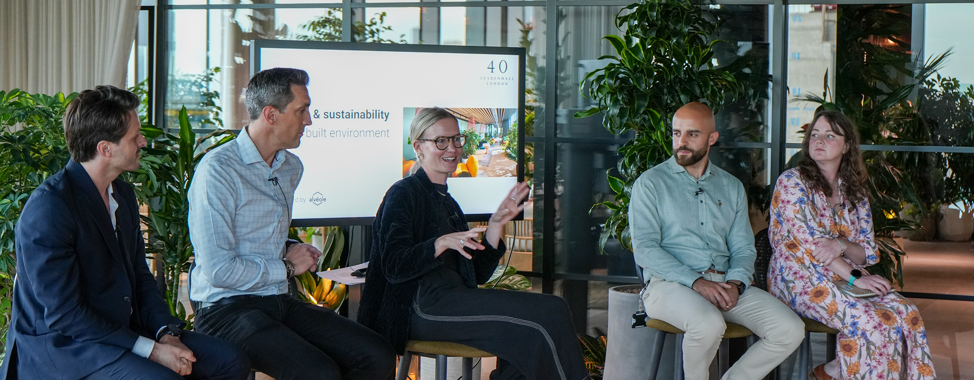 Five panelists sit in a row during a discussion on sustainability in the built environment at 40 Leadenhall London. The woman in the center is speaking animatedly with hand gestures, while the others listen attentively. A screen behind them displays the event topic. The setting includes lush green plants and floor-to-ceiling windows.