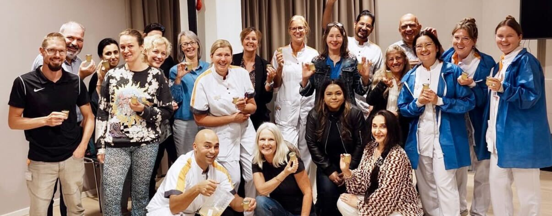 Group photo of about 20 people smiling indoors, many in medical uniforms or blue lab coats, holding small honey-tasting cups. Two people in front crouch down holding honey-related items. The group appears to be celebrating or participating in a tasting session.